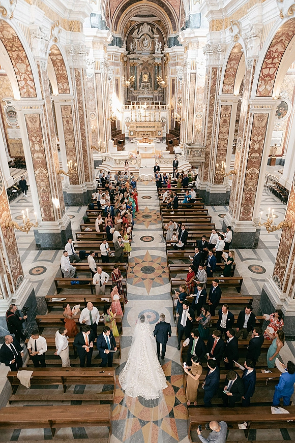 Wedding processional with bride walking down aisle in a bridal gown and long veil through an ornate church with chandeliers and candles