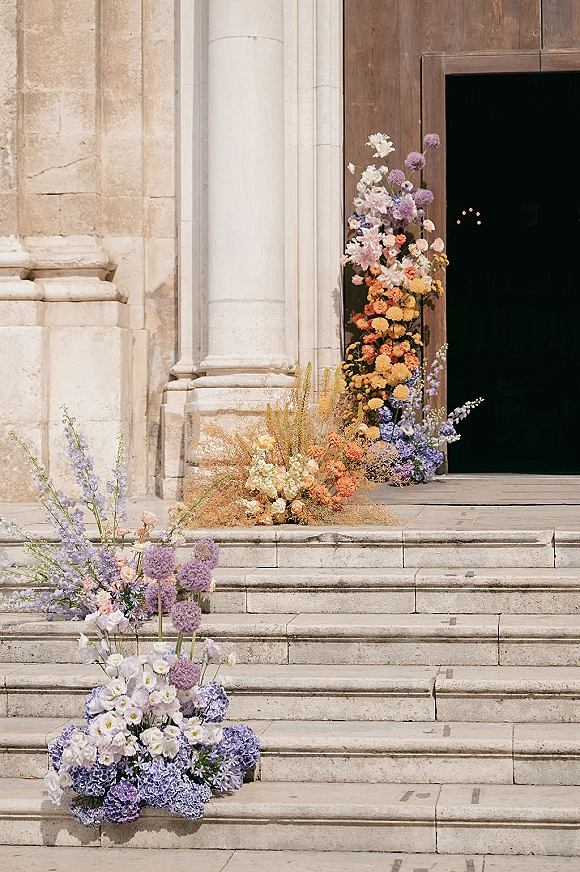Ceremony entrance florals frame the church doorway with pastel hydrangeas, delphinium, and allium on stone steps, accented by meadow blooms