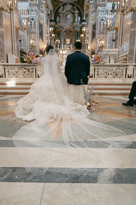 Ceremony moment with bride and groom kneeling at the altar, her cathedral veil and lace train flowing in an ornate church with chandeliers