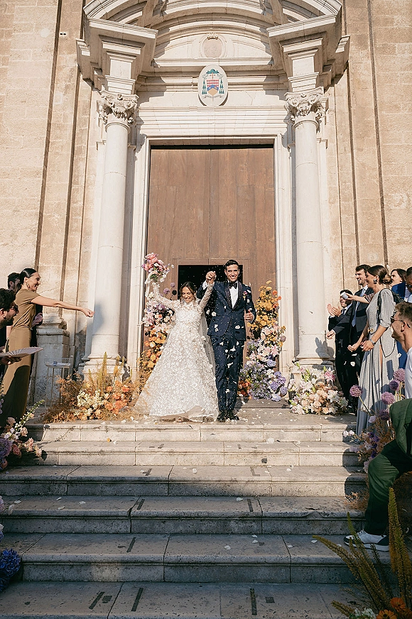 Wedding recessional as newlyweds walk out of a stone church doorway, bride in veil with bouquet, guests tossing flower petals on steps
