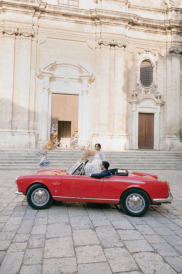 Wedding couple portrait in a red vintage convertible, bride in lace gown and groom in navy suit on stone church steps by arched doorway