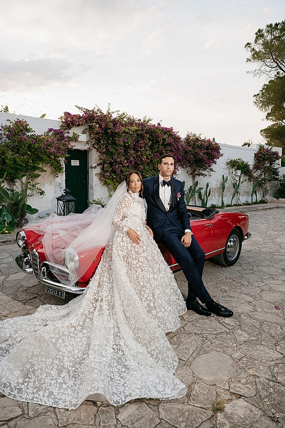 Couple portrait of bride in lace gown with cathedral veil and groom in tuxedo beside a vintage convertible in a stone courtyard