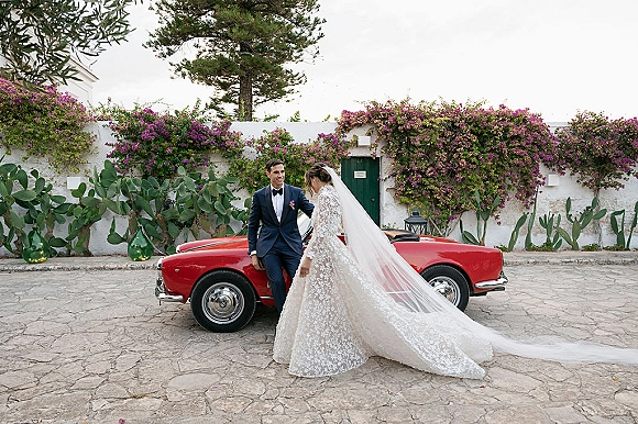 Couple portrait of bride in a long veil and lace dress with groom by a vintage convertible in a stone courtyard with vines and green door
