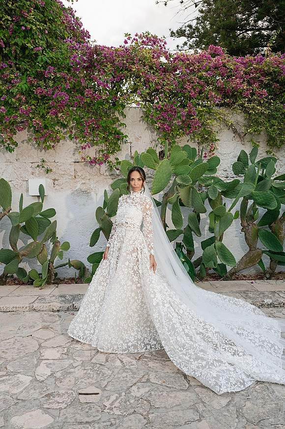 Bridal portrait of a bride in a lace wedding dress with cathedral veil and long train, standing in a stone courtyard with bougainvillea vines