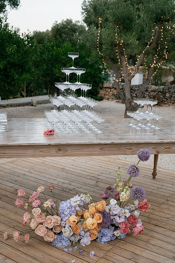 Champagne tower setup with coupe glass pyramid on a wooden table, framed by florals and string lights on an outdoor deck by stone wall