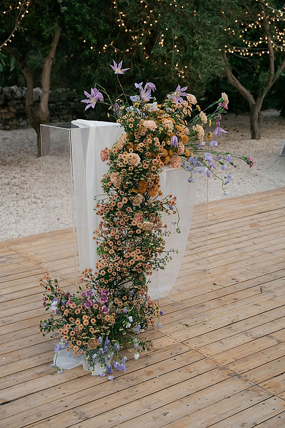 Ceremony altar decor with asymmetrical altar flowers on a clear acrylic pedestal, framed by white drape and string lights on a wood platform outdoors