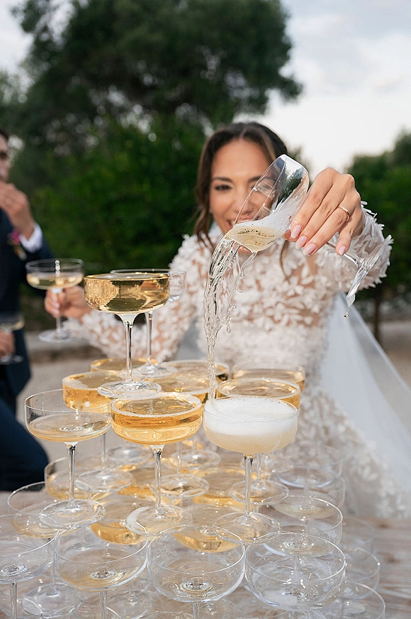 Champagne tower wedding moment as sparkling wine cascades through coupe glasses, framed by bride’s lace sleeve and outdoor greenery behind