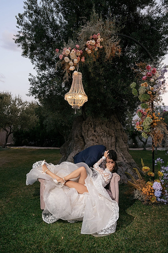Wedding kiss portrait of bride in lace sleeves and veil dipping into groom in navy suit on velvet sofa beneath floral arch and chandelier on garden lawn