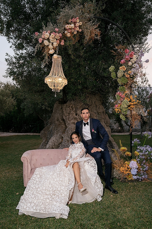 Couple portrait of bride seated on a pink velvet sofa and groom beside her under a chandelier and floral arch at dusk on a garden lawn