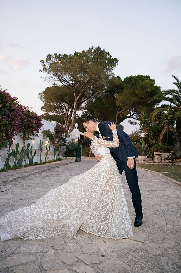 Wedding kiss portrait of bride and groom in a dip kiss wedding photo, her lace long-sleeve gown train sweeping a stone courtyard path.