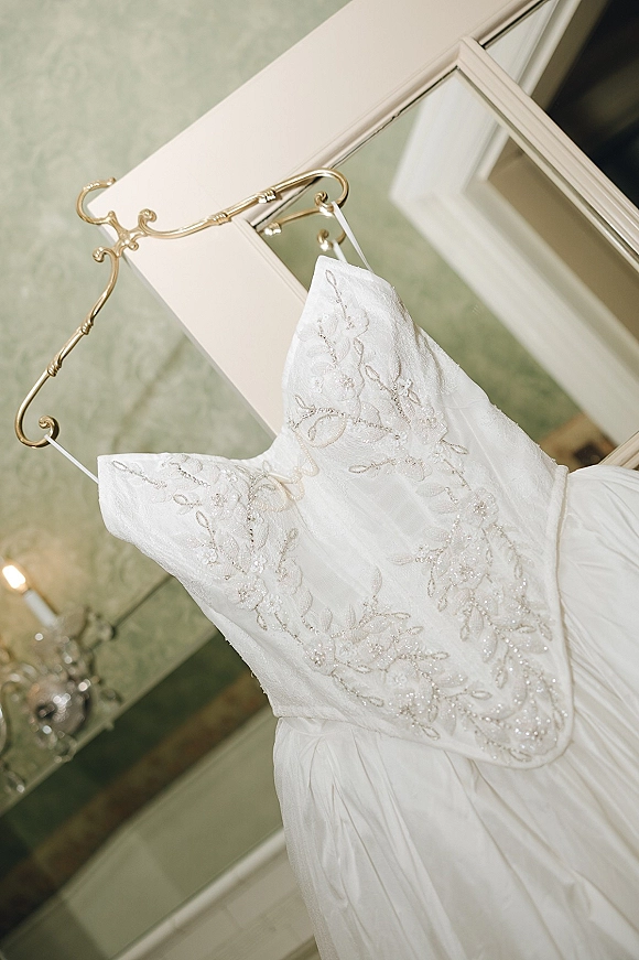 Wedding dress, strapless wedding dress with beaded floral lace bodice and satin waistband on an ornate gold hanger by a mirror in a bridal suite