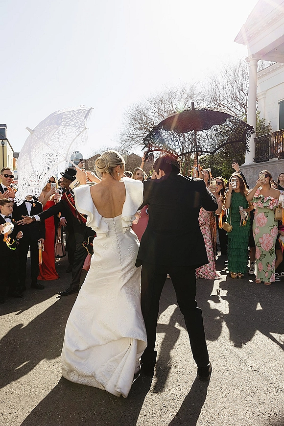 Wedding recessional as bride and groom exit down a sunny street, she holds a lace parasol and bouquet while guests cheer and film behind