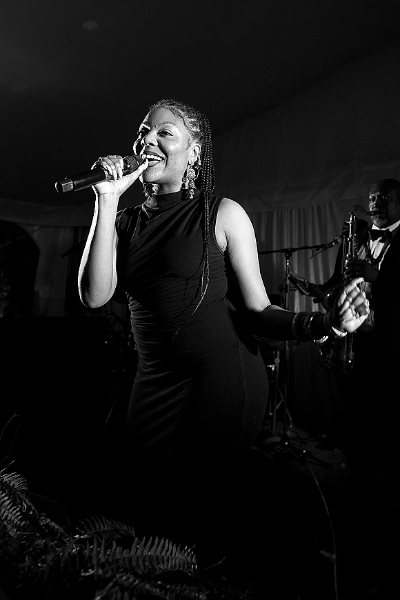 Wedding band singer performing with a handheld microphone in a black dress on stage, with saxophonist and draped backdrop under lights