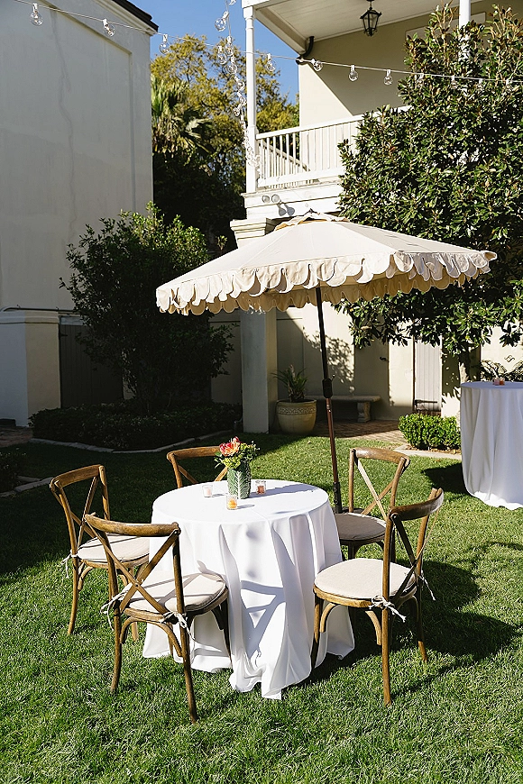 Outdoor cocktail tables with white linens and patio umbrellas, bud vase flowers and votive candles on a lawn with string lights near a white estate porch