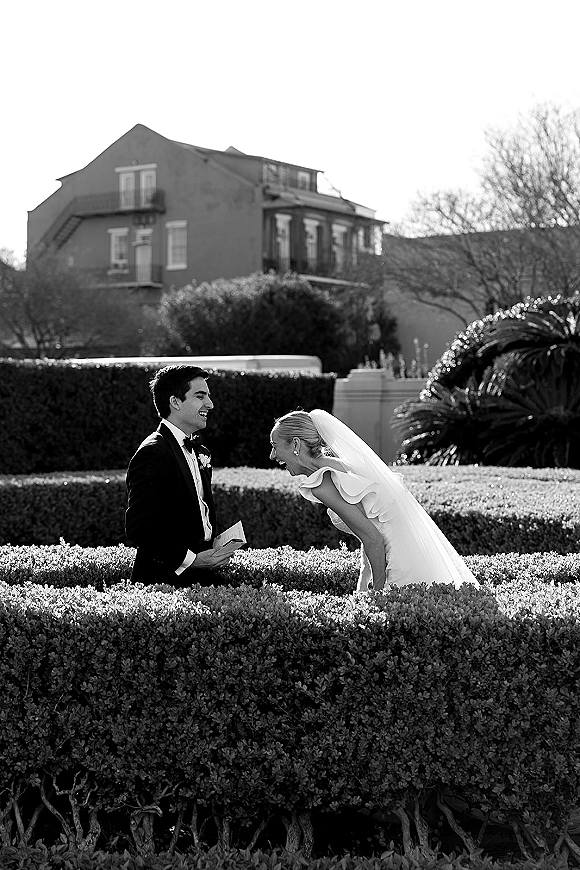 Wedding first look as bride and groom laugh together, veil blowing back over strapless dress, groom in tux with boutonniere in hedged garden