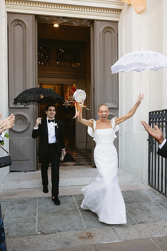 Wedding recessional as bride and groom exit a church doorway, bride cheering with bouquet ribbons while holding white and black parasols on stone steps