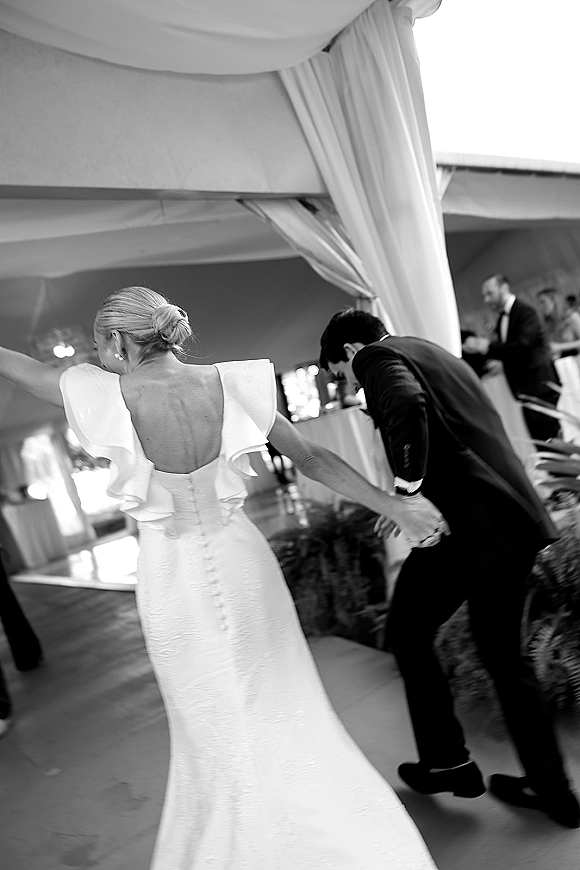 Reception dancing as the bride leads the groom hand in hand on a tent dance floor, her button-back dress under draped canopy lighting