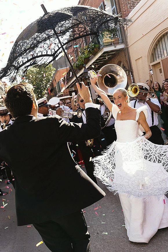 Wedding second line couple dancing in the street as a second line parade follows, bride with lace parasol amid confetti and brass band on brick-lined block