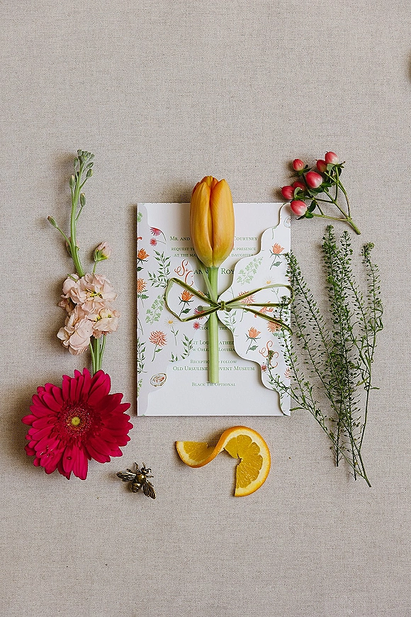 Invitation suite wedding invitation flat lay with floral scalloped card, yellow tulip, green ribbon bow, orange slice, and bee pin on linen backdrop
