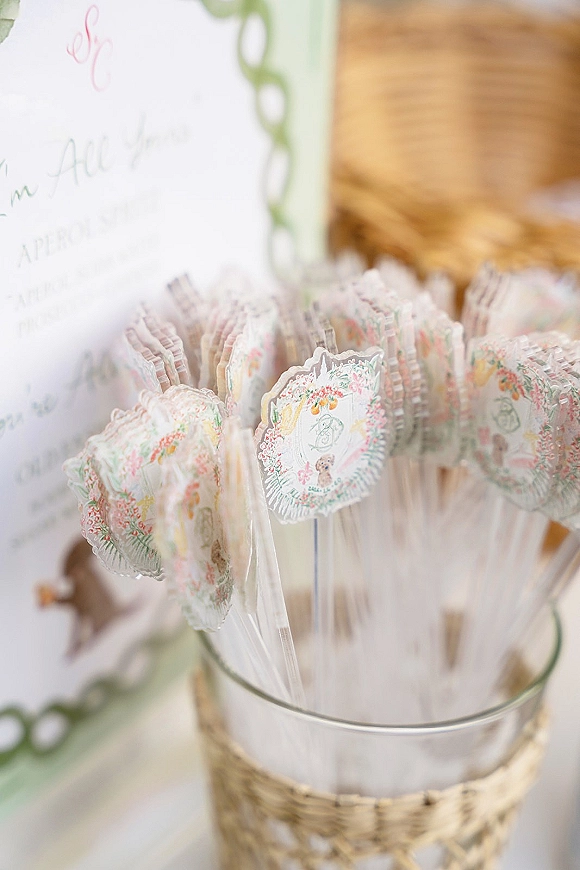Wedding program fans with monogram print and floral illustration on clear sticks, displayed in a glass container on a wood surface