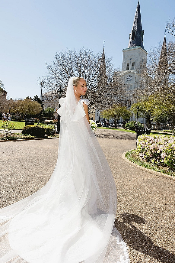 Bridal portrait from behind in a back view wedding dress, long cathedral veil blowing on a garden path with a church steeple ahead