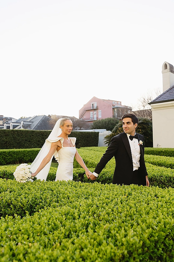 Couple portrait of bride and groom holding hands, her cathedral veil and white bouquet flowing as they stroll by manicured garden hedges