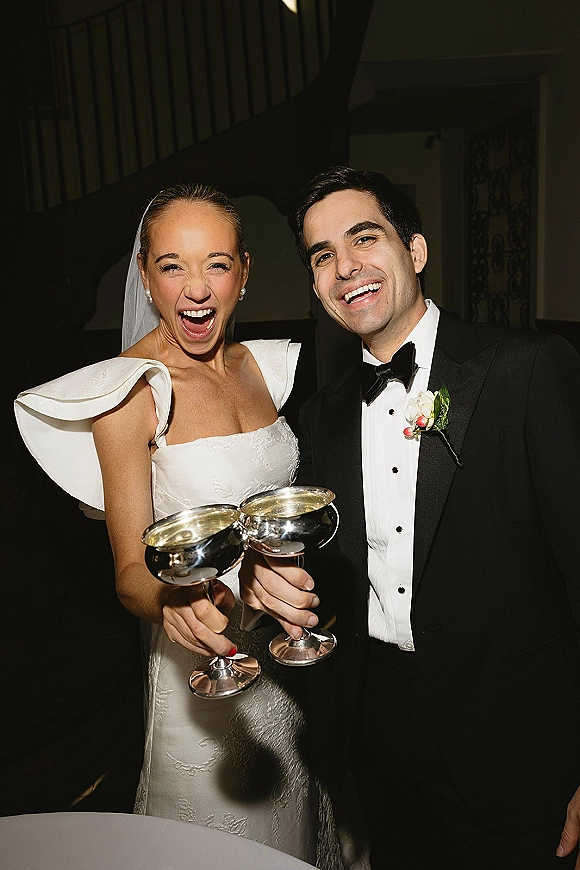 Wedding couple toast with coupe glasses, bride in satin dress and veil laughing beside groom in black tux by an indoor staircase