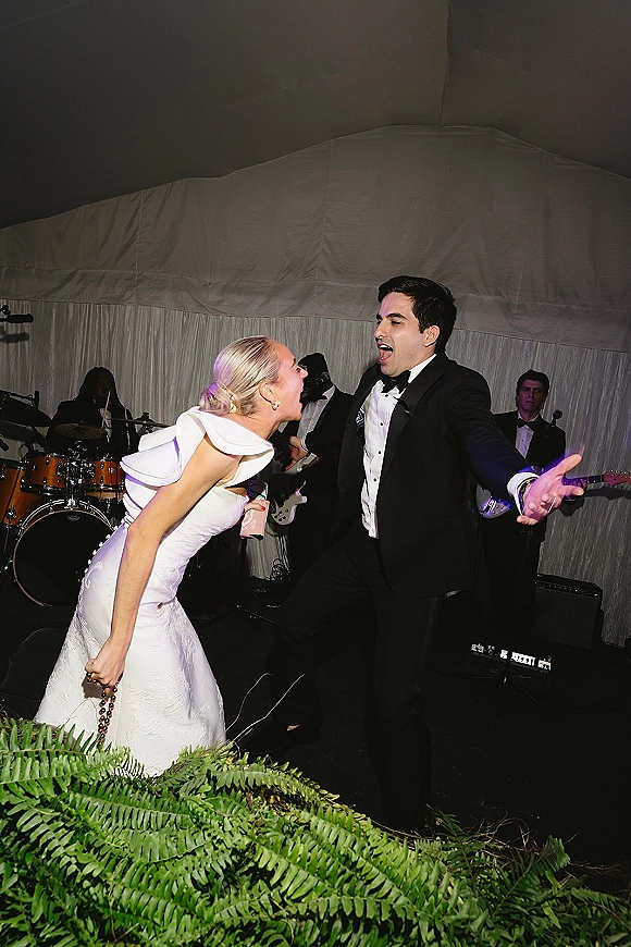 Wedding reception dancing as bride in off-shoulder dress and groom in tux sing along, cocktail cup raised, live band on stage in white tent