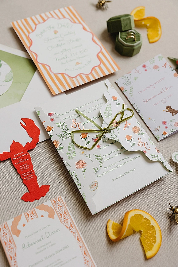 Wedding invitation suite with floral wedding invitations, olive green ribbon, wax seal, rings in velvet box, and orange slices on linen