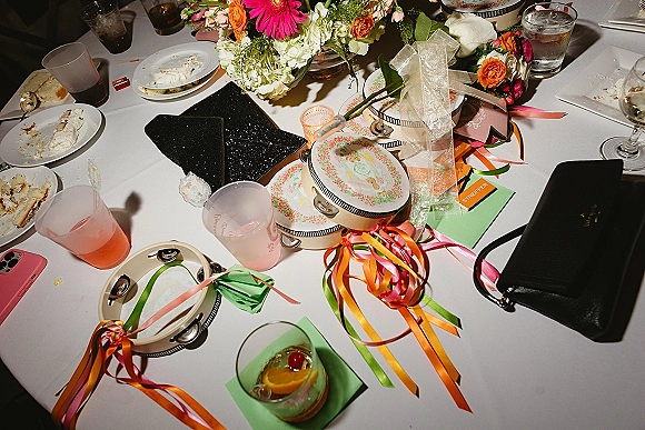 Reception tablescape with wedding table centerpieces—bright floral centerpiece, tambourines, ribbon streamers, cocktails, and votive candles on white linen.