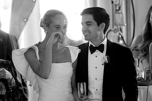 Wedding toast moment as bride wipes tears while groom smiles, both holding champagne flutes at an indoor reception with guests behind