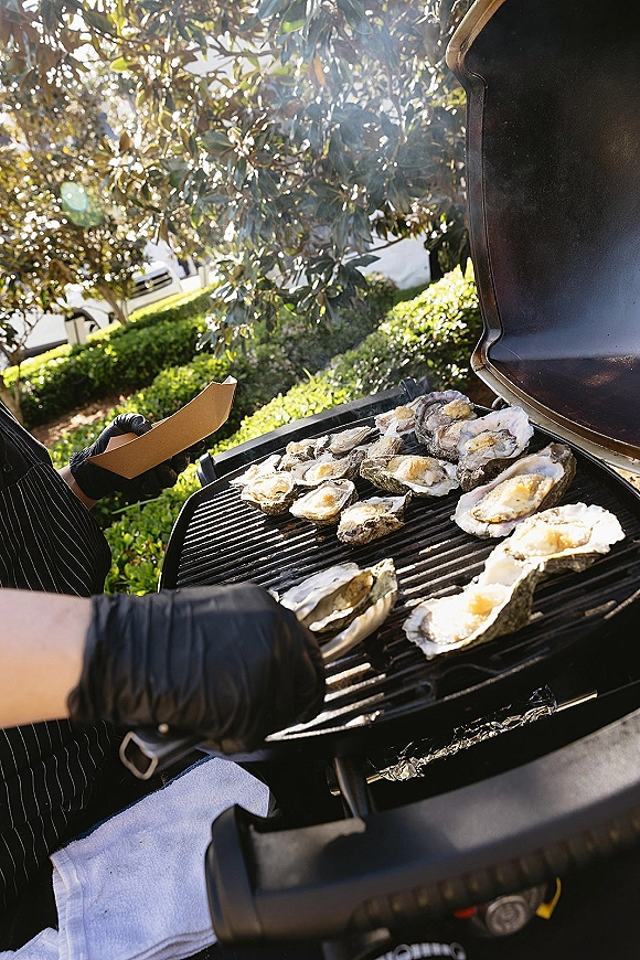 Oyster grill station with chef in black gloves using tongs to cook oysters on foil, set on a sunny lawn with trees and hedges