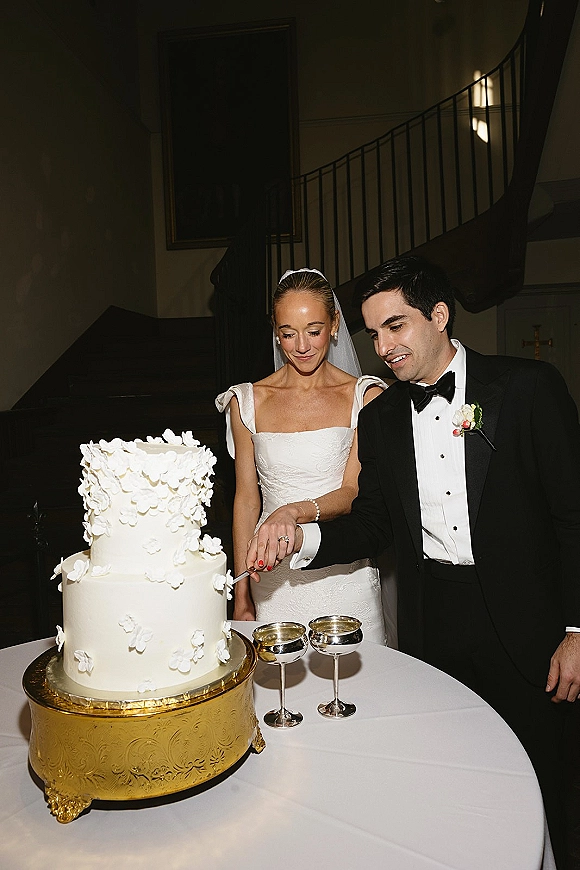 Cake cutting as bride and groom cutting cake on a gold cake stand, veil and tuxedo visible beside coupe glasses in dim indoor reception setting