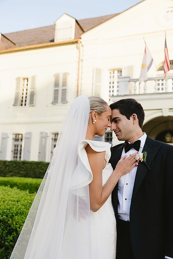 Couple portrait of bride and groom close up, touching foreheads as she holds his lapel; veil and tuxedo before a white estate facade