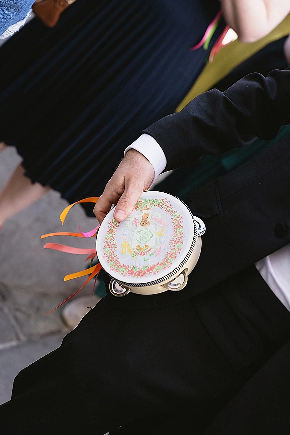 Wedding tambourine with embroidered design and colorful ribbon streamers held by someone in a black suit, with pavement and people behind