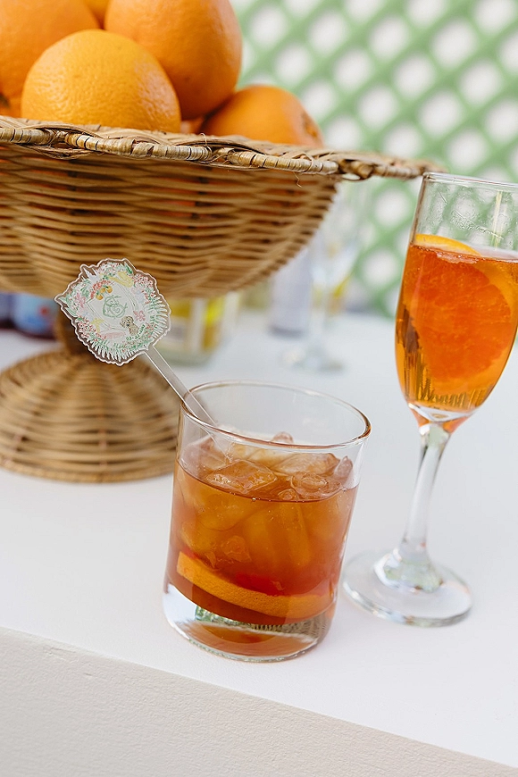 Wedding signature drink with iced cocktail glasses and orange slice garnish, styled on a white tablecloth with green patterned backdrop.