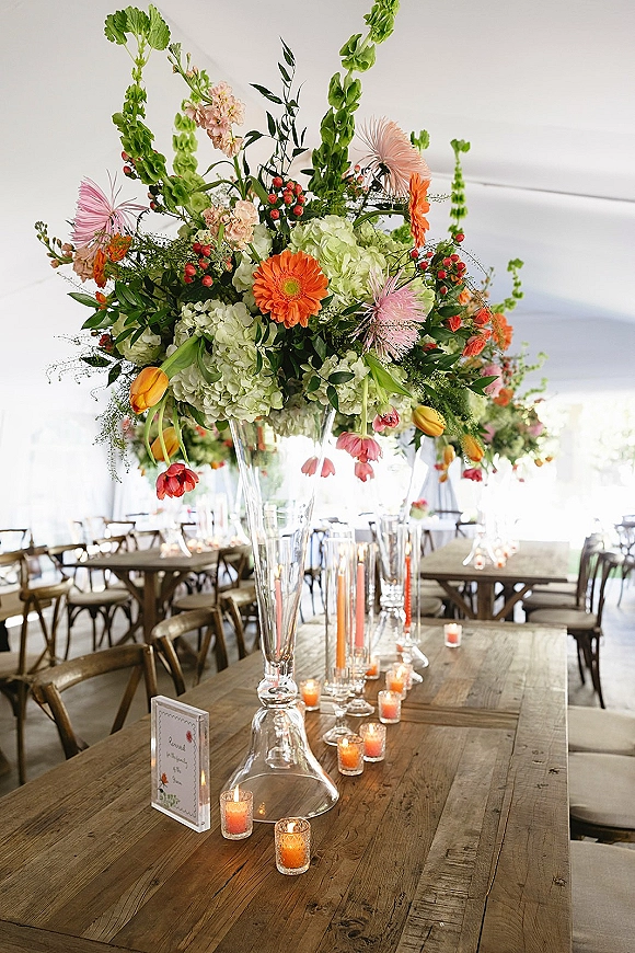 Reception tablescape with a tall floral centerpiece in a glass vase, colorful blooms and votive candles on rustic wood farm tables under a tented ceiling