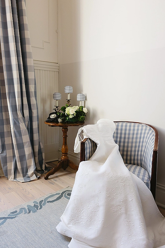 Wedding dress detail with lace wedding dress embroidery draped over an upholstered armchair beside a side table with roses and a vintage telephone