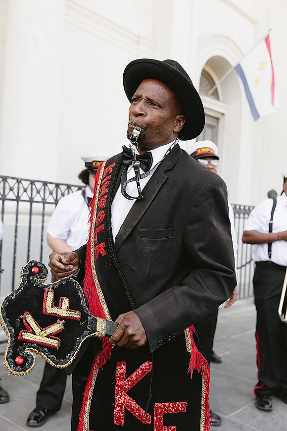 Wedding second line grand marshal in a black suit and bow tie blows a whistle, leading marching band past a white arched facade with flags