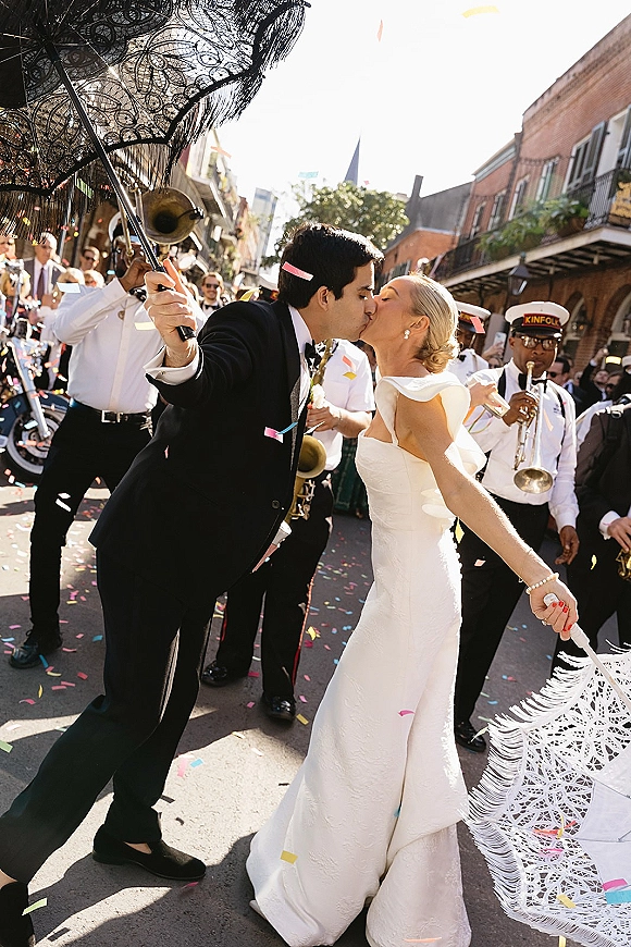 Wedding kiss moment as newlyweds kiss in street under a lace parasol while confetti falls, with brass band and crowd along brick balconies