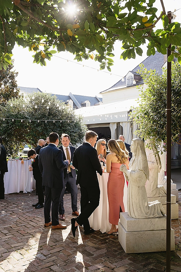 Wedding cocktail hour with outdoor cocktail hour guests mingling by white linen high-top tables under string lights in a sunny brick courtyard