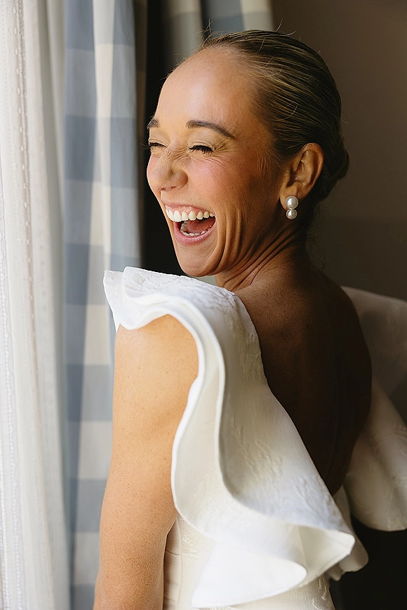 Bridal portrait of a laughing bride looking over her shoulder in a wedding dress, wearing pearl drop earrings by a window with curtains