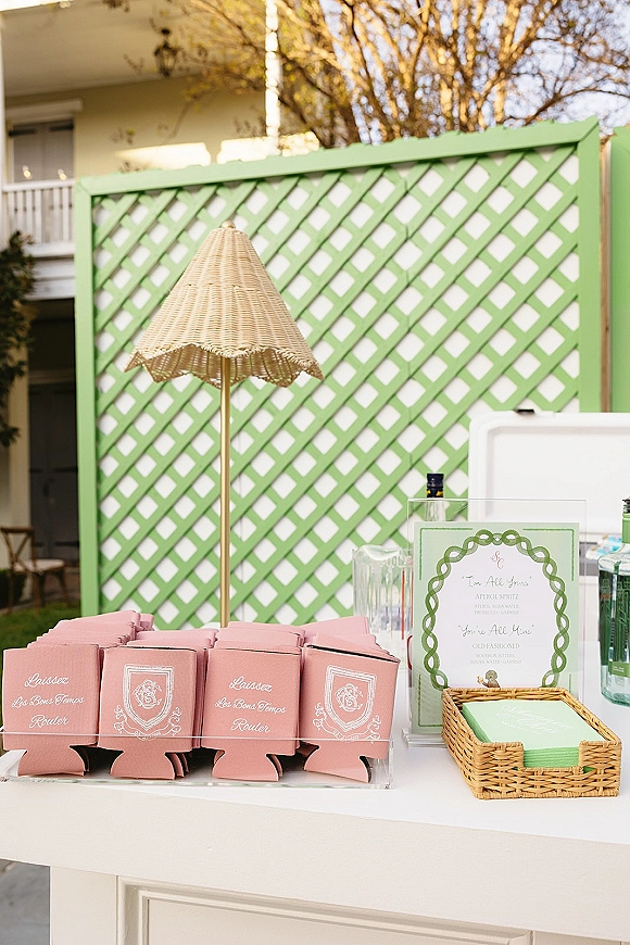 Wedding bar setup with wedding cocktail station drink menu sign, glassware and bottles on a table with umbrella against green lattice wall