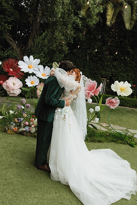 Wedding kiss as bride and groom kissing beneath oversized paper flowers, her long veil and embroidered gown train on a garden lawn with string lights