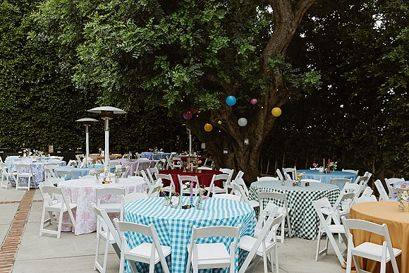 Outdoor reception setup with round tables in patterned tablecloths, bud vases and votive candles under string lights by a large tree canopy