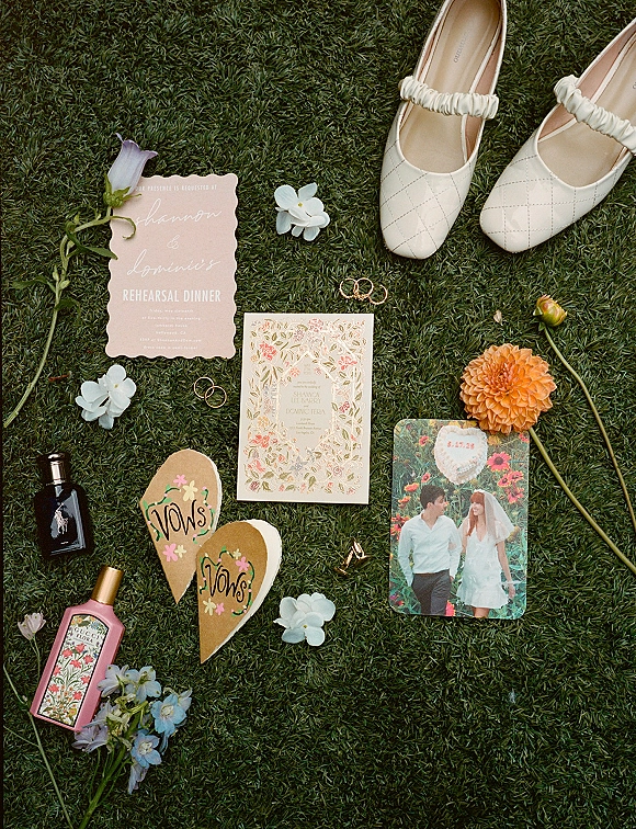 Wedding flat lay with wedding invitation suite, rings, white Mary Jane bridal shoes, perfume, vow cookies, flowers arranged on grass