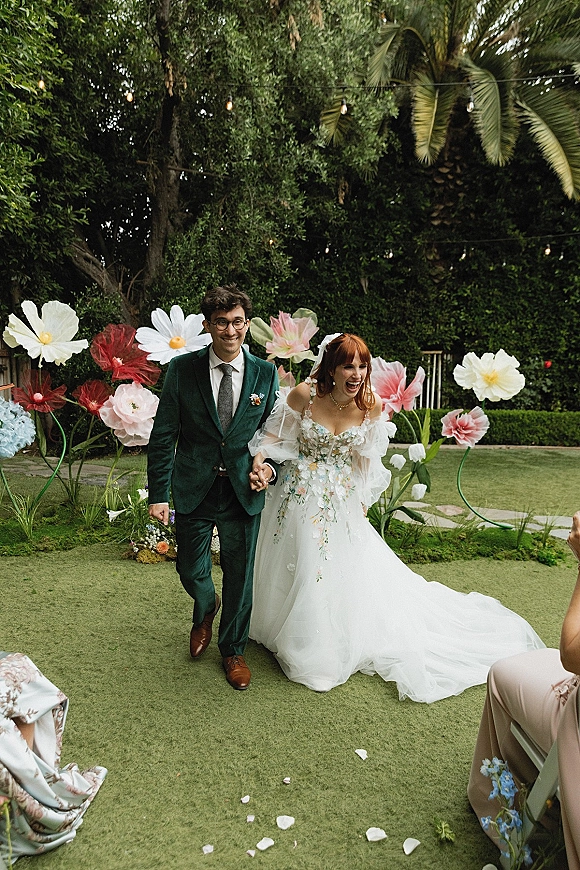 Recessional moment as bride and groom walking hand in hand, smiling under string lights with oversized paper flowers on a garden lawn aisle of petals