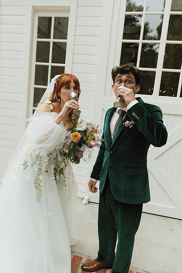 Couple portrait of bride and groom drinking from silver goblets, bride holding bouquet and veil bow before white barn doors