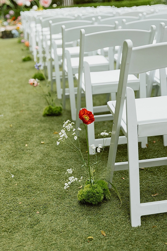 Ceremony aisle decor with outdoor ceremony seating, white folding chairs lined with baby's breath, red poppy, and moss on a garden lawn