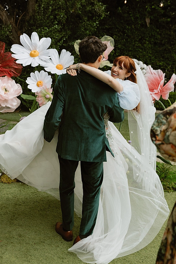 Couple portrait of bride and groom embrace, groom holding a laughing bride in wedding dress and long veil with oversized paper flowers on lawn